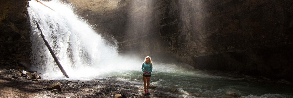 Wahrnehmung verstehen - eine Frau steht auf einem Felsen nahe einem Wasserfall und Sonnenstrahlen fallen von oben auf sie herab